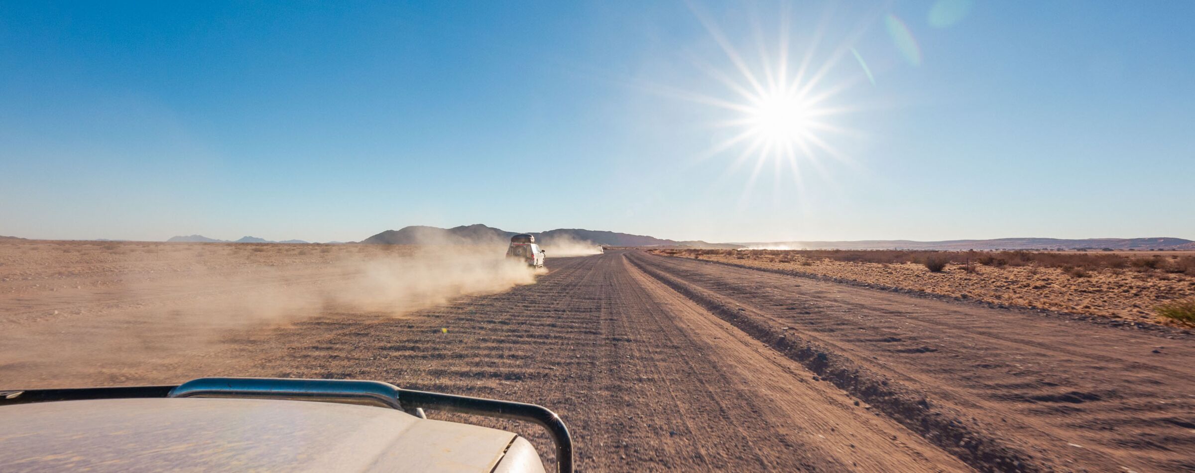 View from an off-road vehicle on a dusty desert road, following a convoy of vehicles under the bright sun.
