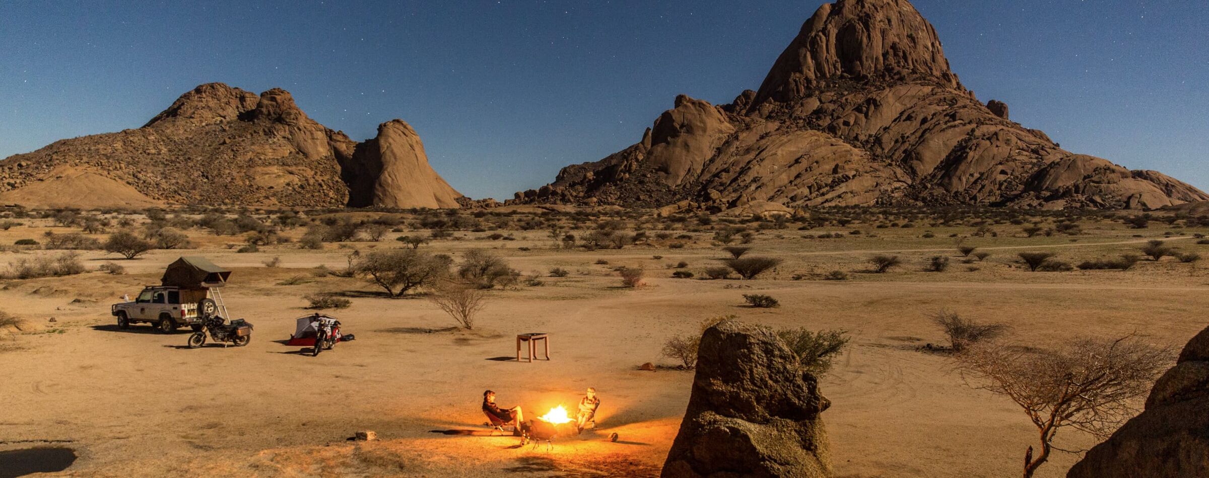Evening atmosphere in Namibia’s desert: Two travelers enjoy a campfire, surrounded by 4x4 vehicles and rocky landscapes – Gravel Travel experience.