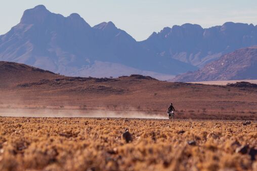 A lone motorcyclist kicks up dust as he rides through the vast African wilderness. Towering mountains in the background add to the sense of adventure and boundless freedom.